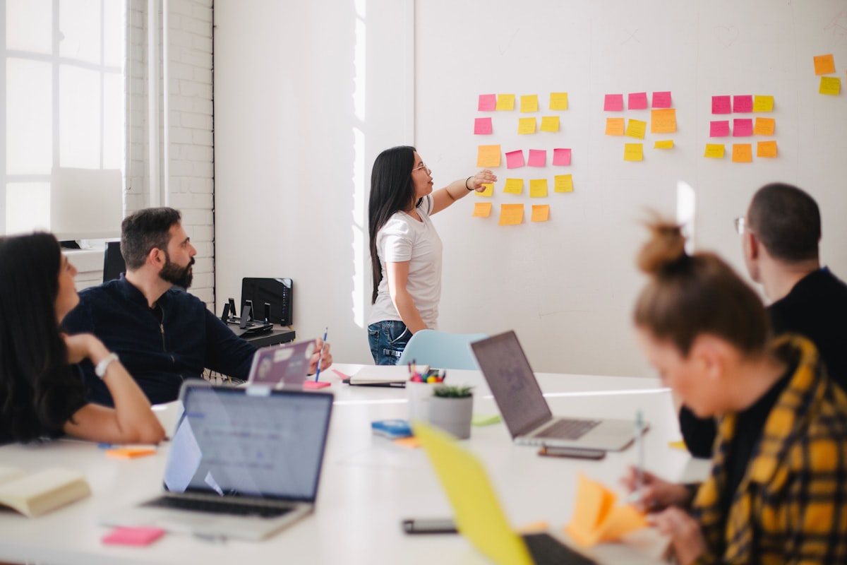 Four people gathered around a wooden conference table brainstorming implementation steps, with sticky notes on a wall behind them.